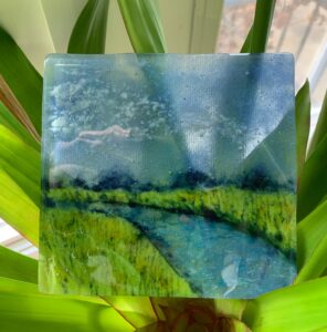 A fused glass piece of a small blue waterway in the middle of a grassy land with a blue and white cloudy sky.