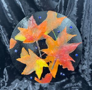 This is an image of fused glass red and yellow maple leaves against a black circular background with dewdrops made of glass.