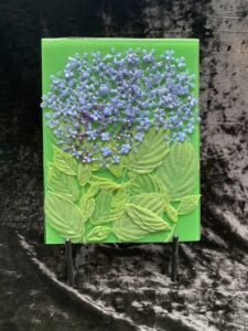 A fused glass piece with powdered glass blue hydrangea flowers and green leaves against a green background.